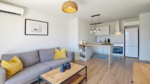 Ein modernes Wohnzimmer mit grauem Sofa, gelben Kissen, einem Holztisch und einer Küchenzeile mit weißen Schränken und einem eingebauten Backofen.