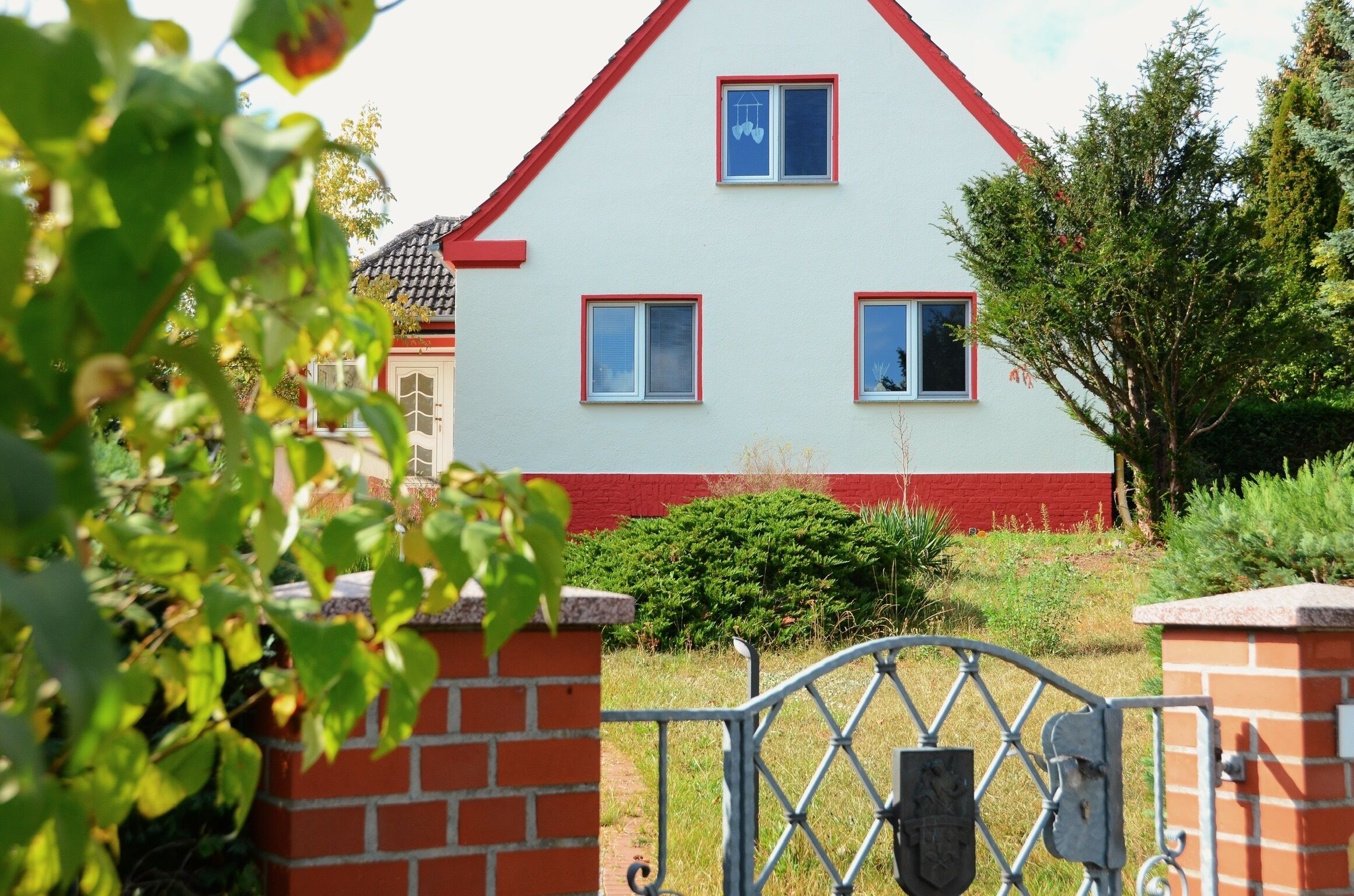A house with a red roof and white walls, surrounded by greenery and a metal gate.