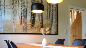 A dining area with a wooden table, black chairs, and two pendant lights. A large forest-themed painting hangs on the wall behind the table. - Ferienhaus zum Glück (Gerswalde)