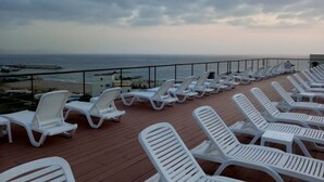 A deck with white lounge chairs facing the ocean, a railing, and a view of the beach and sea. - Duplex In Siwar,2 Minutes From Hotel Le Royal,1 Bedroom, Pool,generator Included (Zouk Mosbeh)