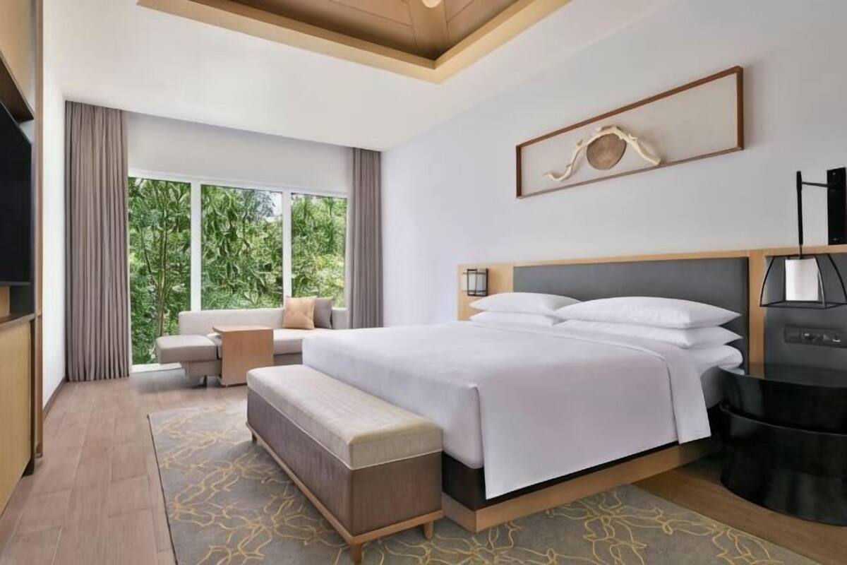 A modern hotel room with a large bed, a seating area, a desk, and a view of the outdoors.