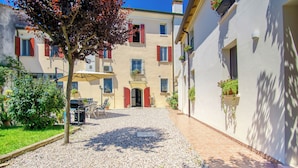 A courtyard with a gravel path, a tree, and a building with red shutters and potted plants.