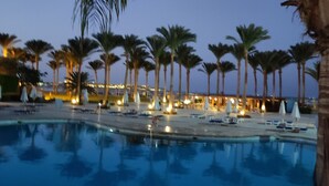 A swimming pool surrounded by palm trees and lounge chairs, with a building in the background. - Chalet Stella Makadi Beach 5 Stars (Makadi Bay)