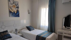 A hotel room with two beds, a desk, a chair, a painting on the wall, and a window with curtains. - Chalet Stella Makadi Beach 5 Stars (Makadi Bay)