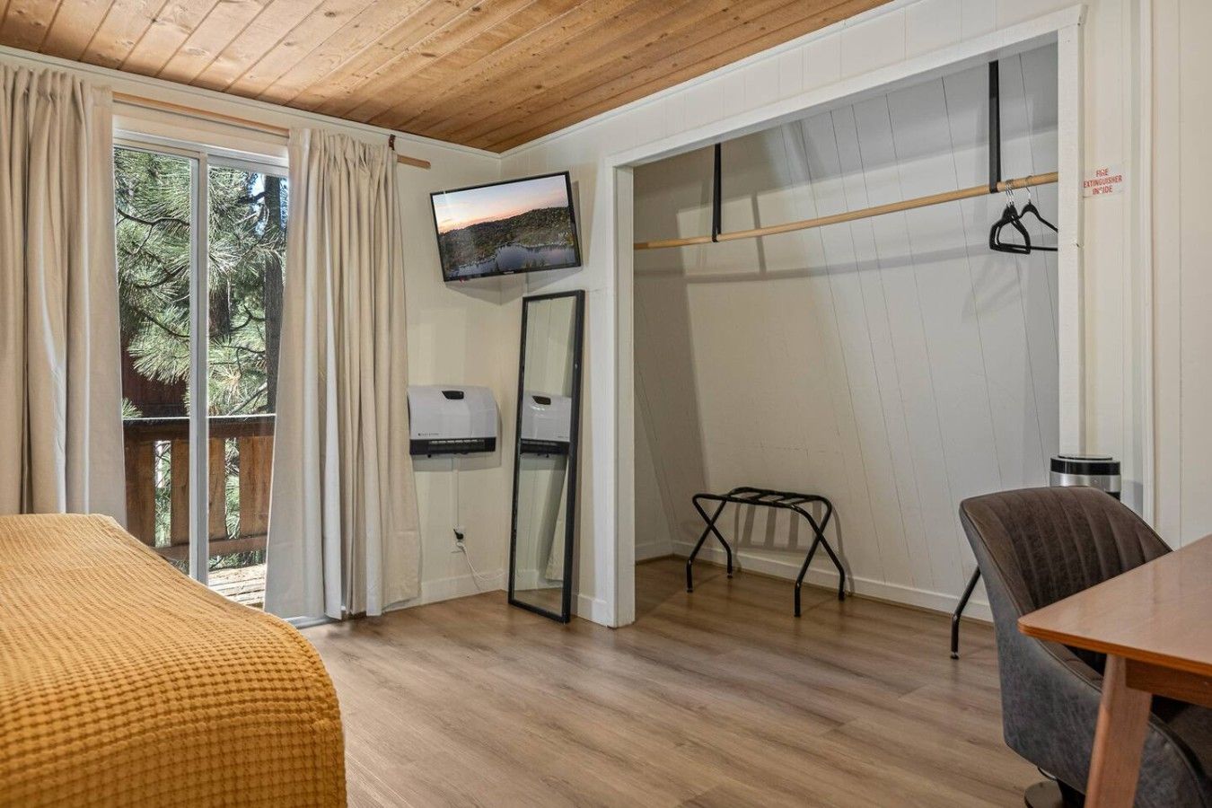 Une pièce comprenant un lit, une table, une chaise, un téléviseur, un porte-manteau et une fenêtre avec des rideaux.