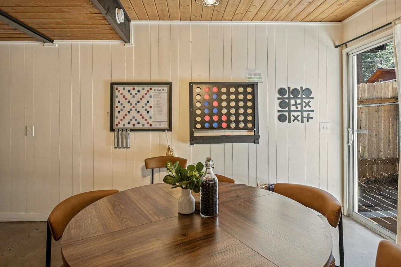 A dining area with a round wooden table, two chairs, a framed Scrabble game, a board game, and a potted plant.