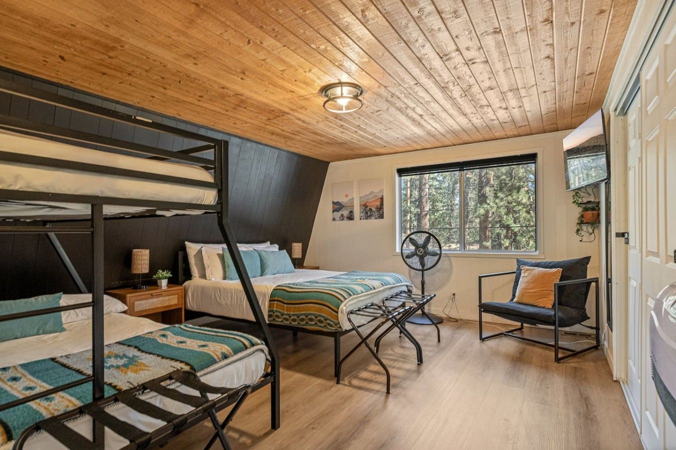 A room with bunk beds, a desk, a chair, a fan, and a window with a view of trees.