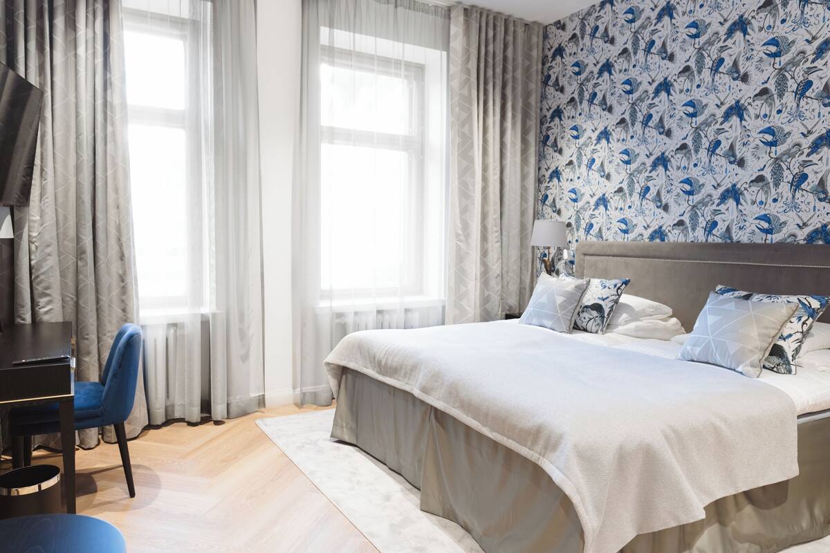 A bedroom with a large bed, a desk, and a chair. There are two windows with sheer curtains and a wall with a blue and white pattern.