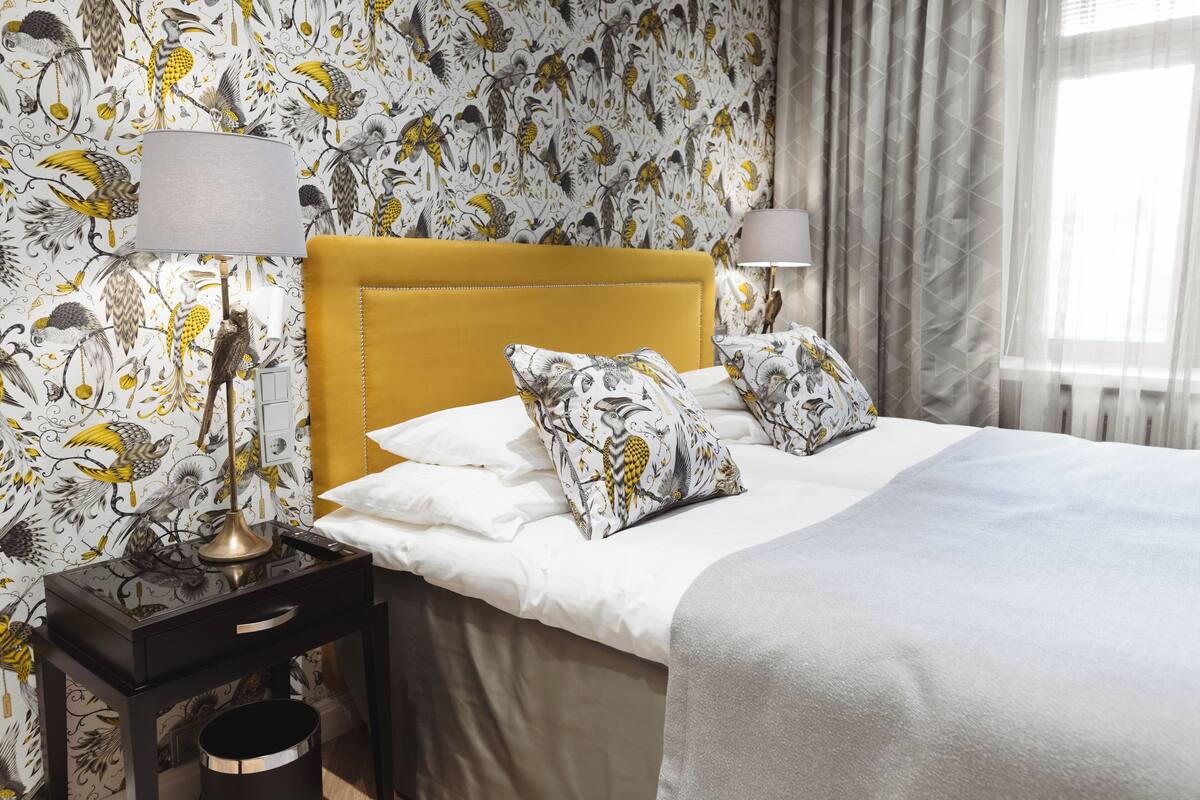 A neatly made bed with white linens and two patterned pillows, a bedside table with a lamp, and a wall with a bird and floral wallpaper.