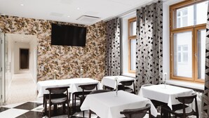 A dining area with tables and chairs, a flat-screen TV mounted on the wall, patterned wallpaper, and a window with blinds. - Hotel Bastian  (Helsinki)