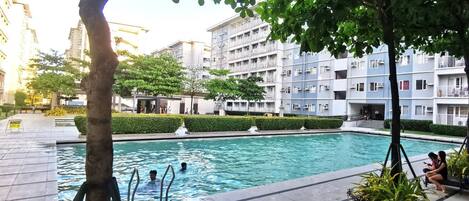 Una piscina rodeada de jardines, con un edificio de varios pisos al fondo.