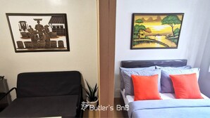 Une chambre d’hôtel avec un lit, une chaise, un tableau représentant un paysage de village accroché au mur et une plante en pot.
