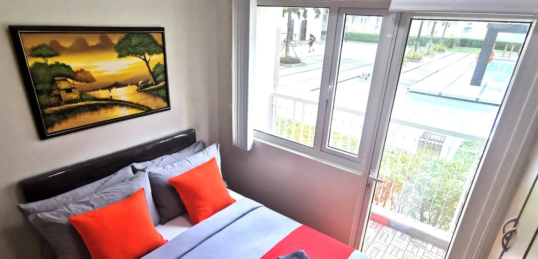 Quarto com cama, almofadas, pintura e janela com vista para uma varanda e área externa.