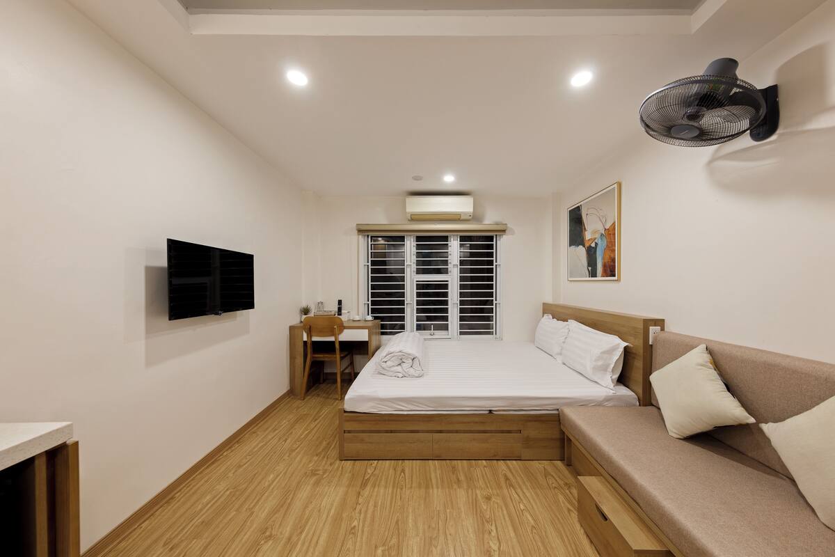 Une chambre à coucher comprenant un lit, un bureau, une chaise, un téléviseur, un ventilateur fixé au mur et une fenêtre avec des stores.