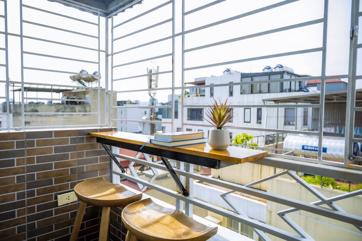 Un balcon donnant sur des immeubles, une plante en pot et un tabouret en bois.