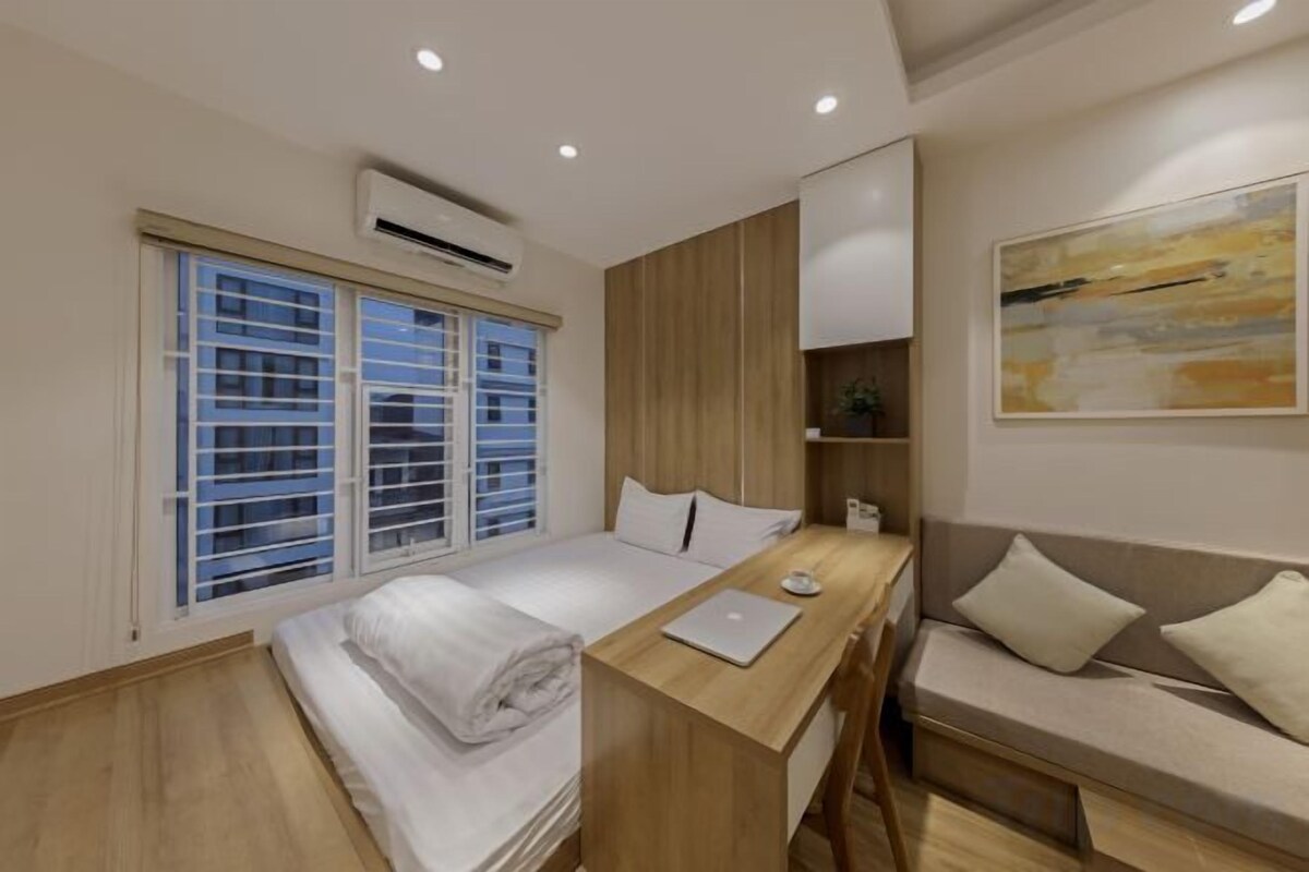 Quarto de hotel moderno com cama, escrivaninha, sofá e uma janela grande com persianas.