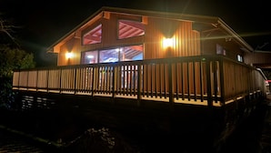 A wooden cabin with a balcony, large windows, and a warm light source inside.