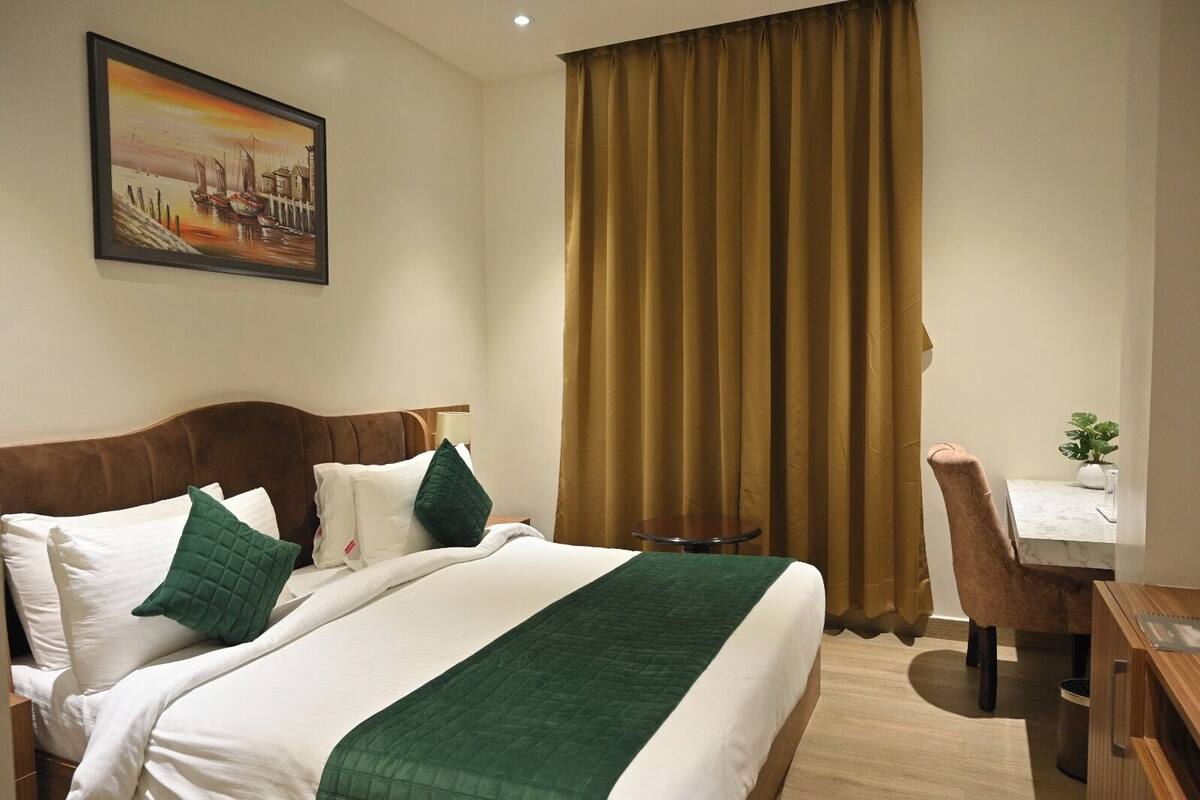 A hotel room with a bed, a desk, a chair, a painting, and a window with curtains.