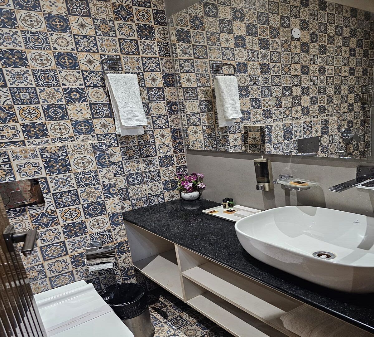 A bathroom with patterned tiles, a white sink, a mirror, and a vase with flowers.