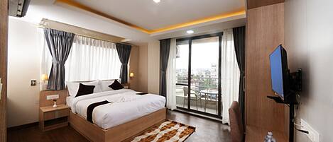 A modern hotel room with a large bed, a flat-screen TV, a balcony with a city view, and a wooden floor.