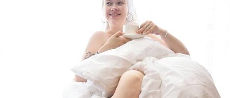 Una persona sentada en una cama con sábana blanca, vestida con una blusa blanca y sosteniendo una taza.