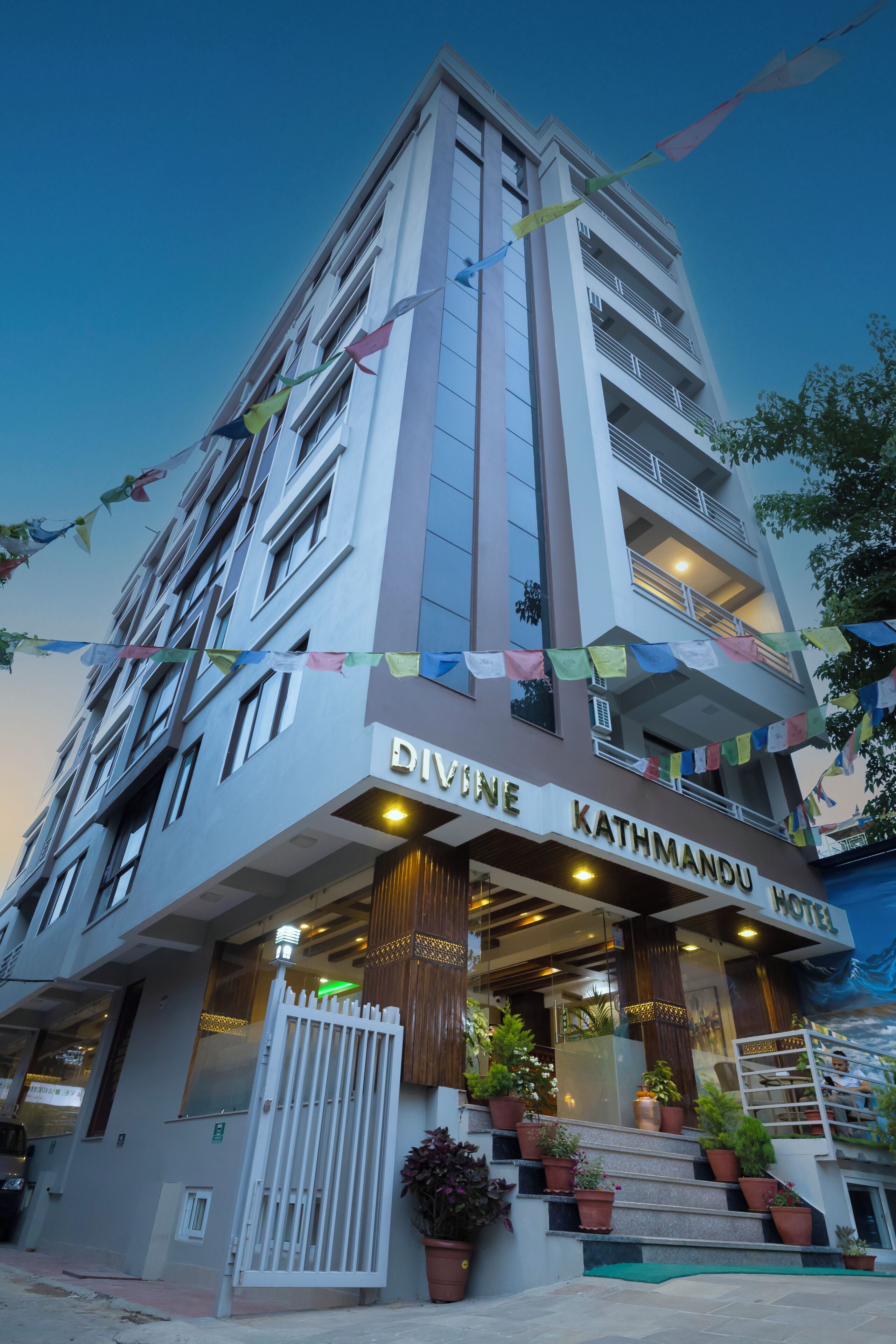 A multi-story building with a sign reading "Divine Kathmandu Hotel." It features a modern design with a glass entrance, potted plants on the steps, and a gated entrance.