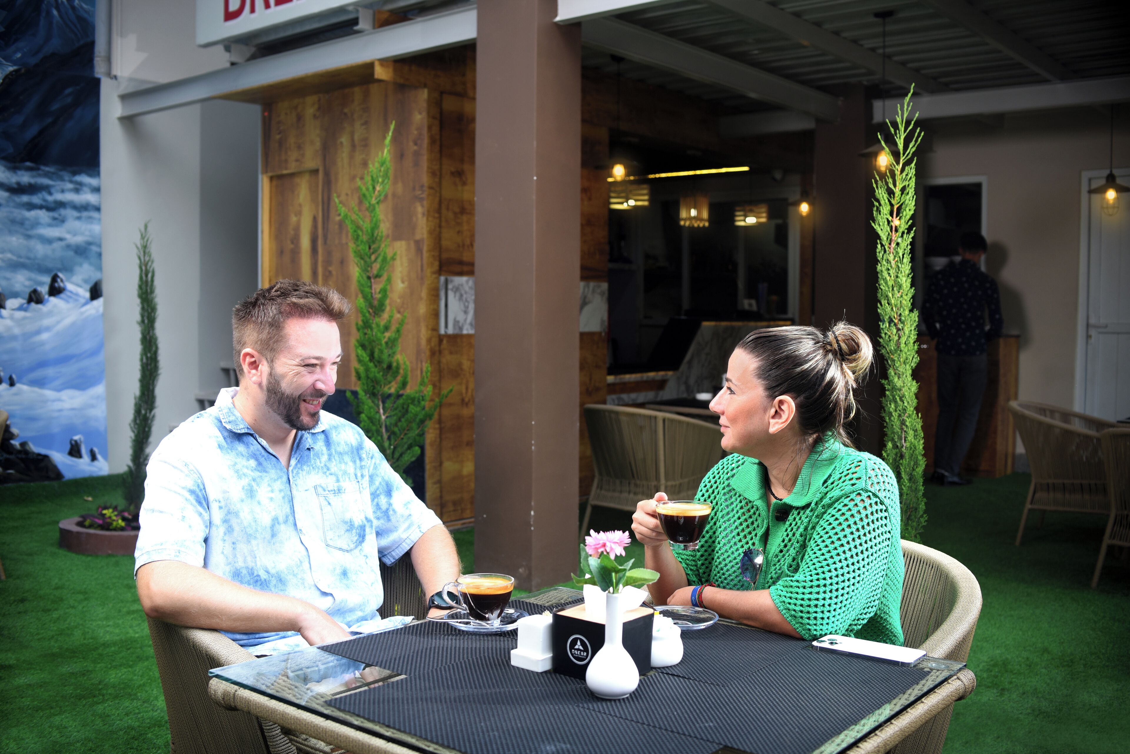 Dos personas sentadas en una mesa al aire libre, con una taza de café y un florero de flores.