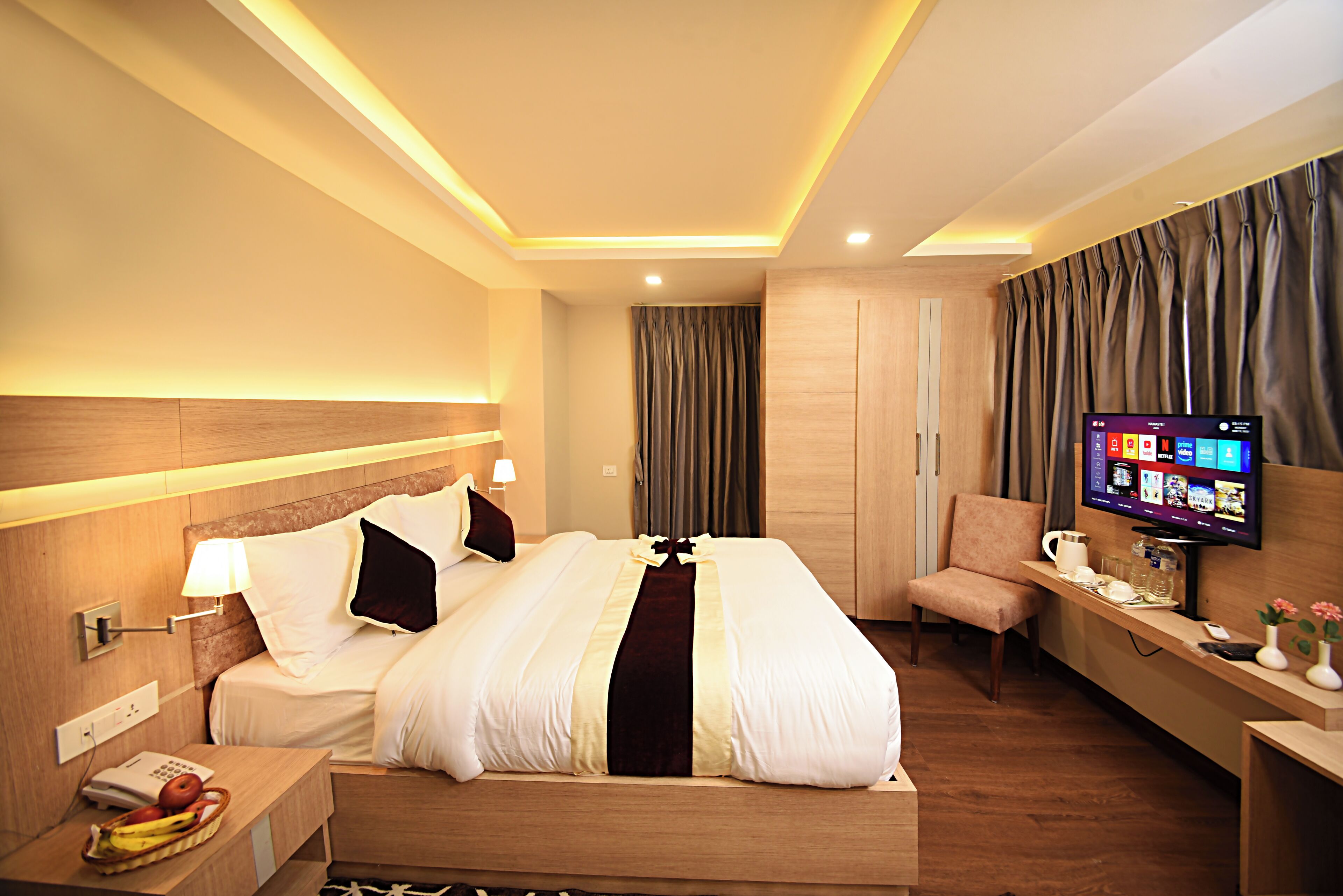 A hotel room with a large bed, bedside table with a lamp, a TV on a stand, a chair, and a desk with a phone and a vase of flowers.