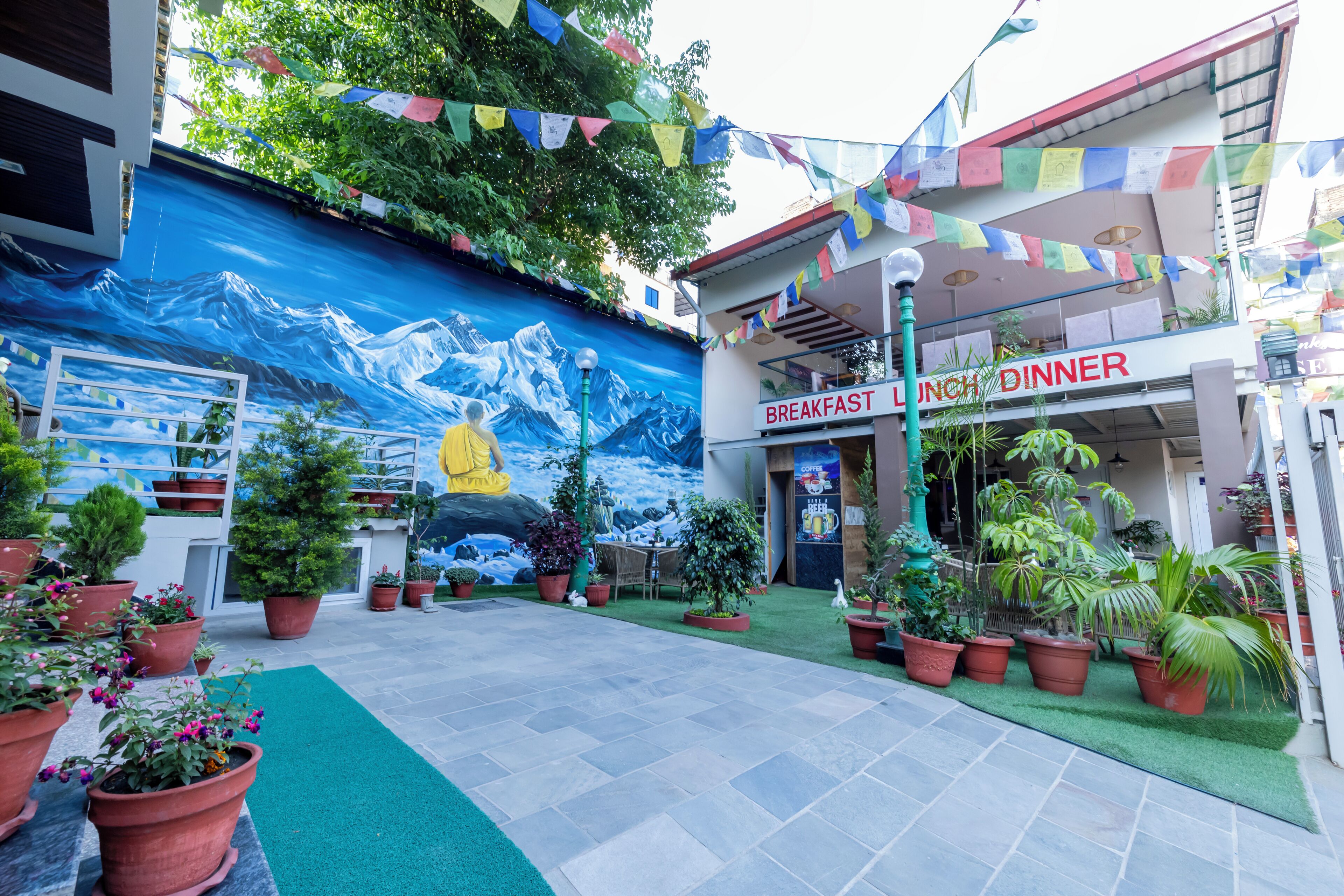 Un pati amb un mural de muntanyes, plantes en testos i un restaurant anomenat "Breakfast Lunch Dinner".
