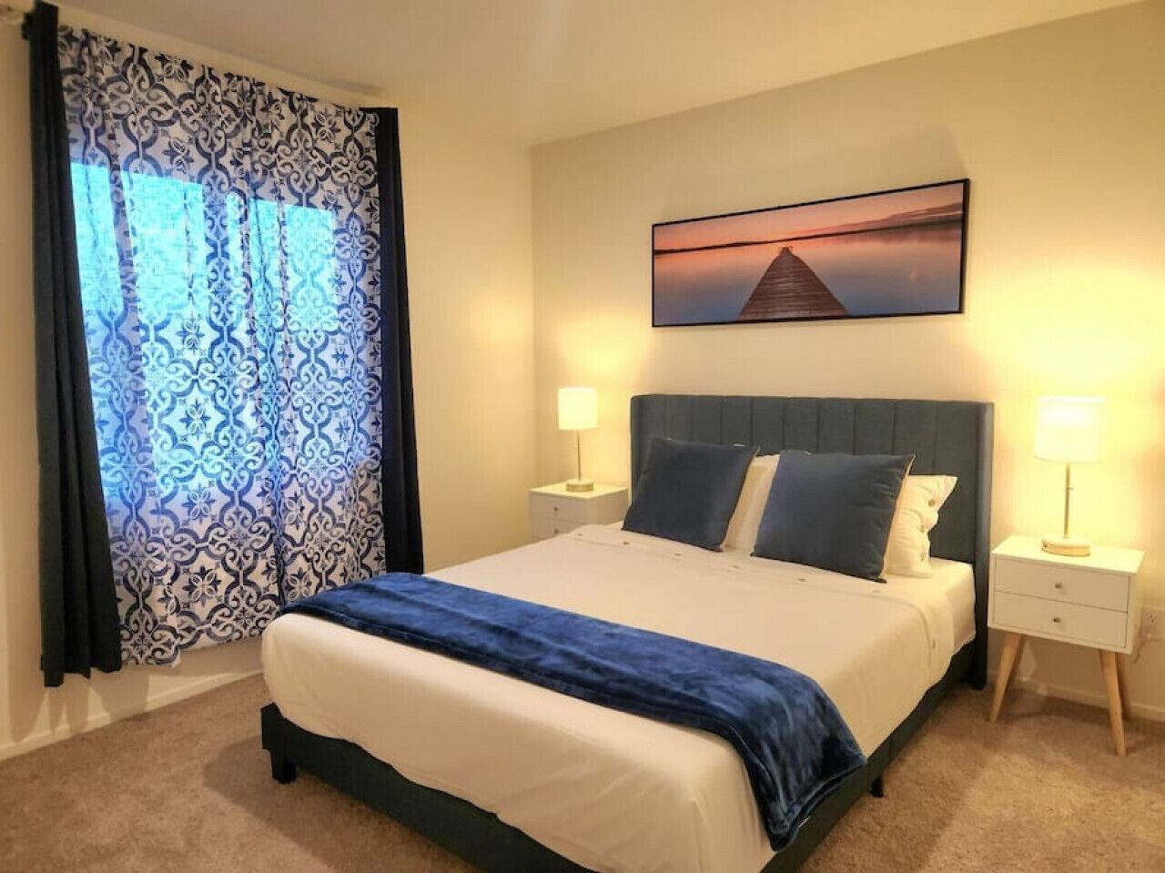 Een net opgemaakt bed met een blauw-witte dekbedovertrek, twee nachtlampjes, een ingelijst schilderij boven het bed, en een raam met gordijnen met een patroon.