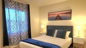 Una cama bien hecha con colcha azul y blanca, dos lámparas de noche, un cuadro enmarcado sobre la cama y una ventana con cortinas estampadas.
