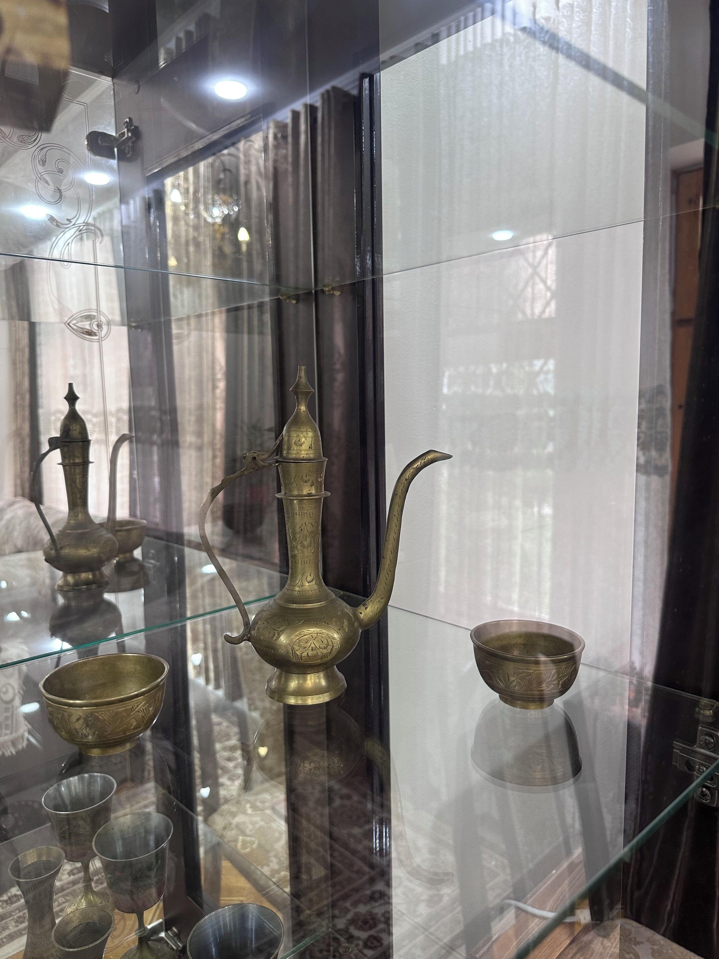 Una exhibición de artefactos de latón, incluyendo una tetera, tazas y un cuenco, sobre una estantería de vidrio.