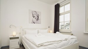 A neatly made double bed with white linens, a bedside table with a lamp, and a window with curtains. - Die Ferienwohnung Nr. 04 im Haus Rugier in Göhren (Göhren)