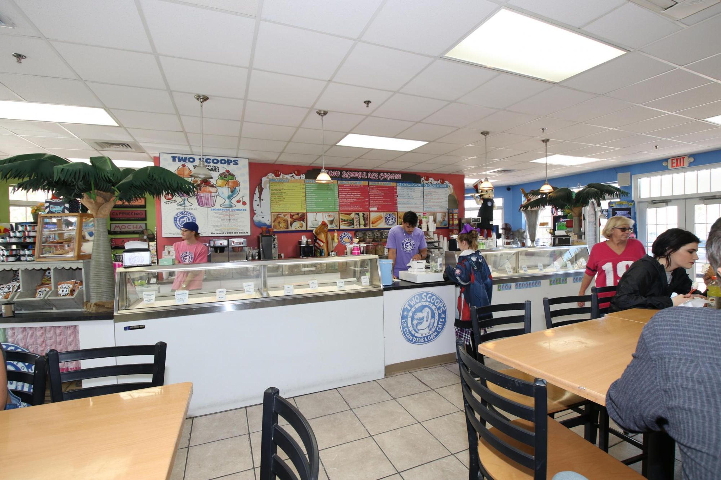 A food court with a counter displaying various food items, a menu board, and a few people dining.
