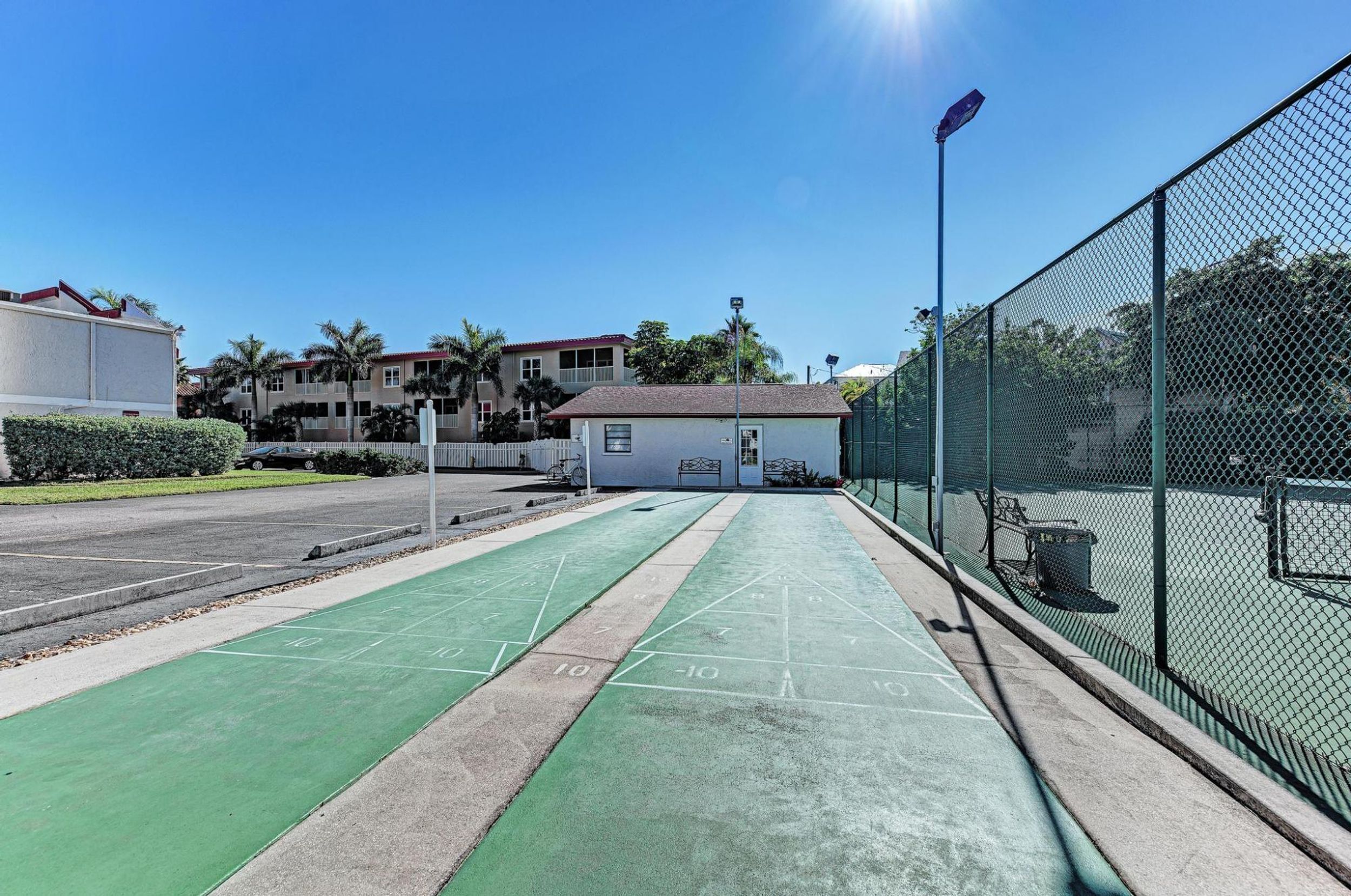 Una cancha de shuffleboard verde al lado de una cancha de tenis, rodeada por una cerca de malla, con un pequeño edificio al fondo.