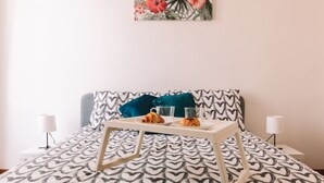 Een keurig opgemaakt bed met een sprei met hartjesprint, een dienblad met croissants en glazen, en een bloemrijk kunstwerk aan de muur.