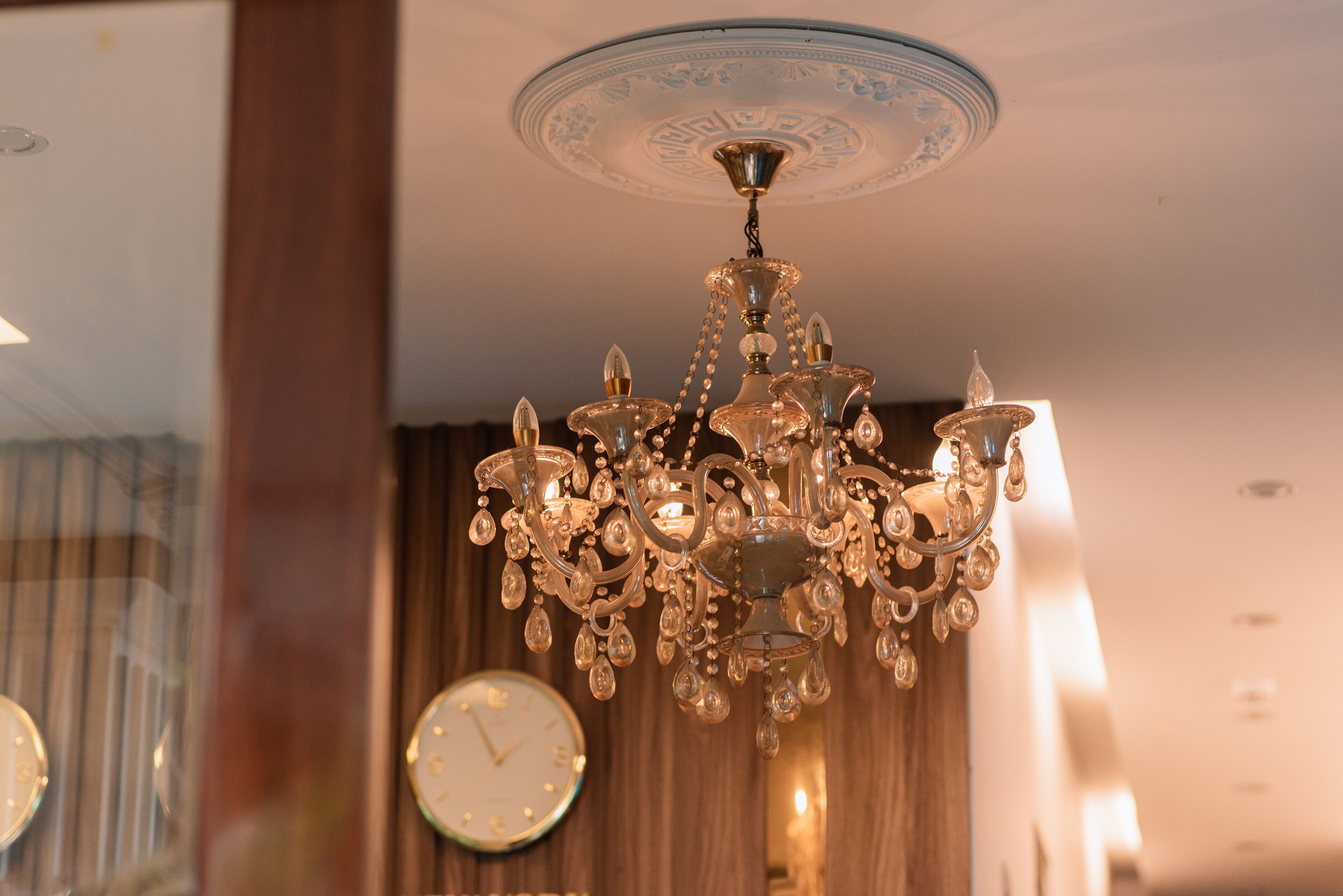 A chandelier with multiple arms and hanging crystals, a clock on the wall, and wooden paneling in the background.