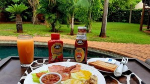Ein Frühstücksbrett mit Eiern, Speck, Bohnen, Toast und einem Glas Orangensaft, mit Gewürzen und Blick auf einen Pool im Hintergrund.