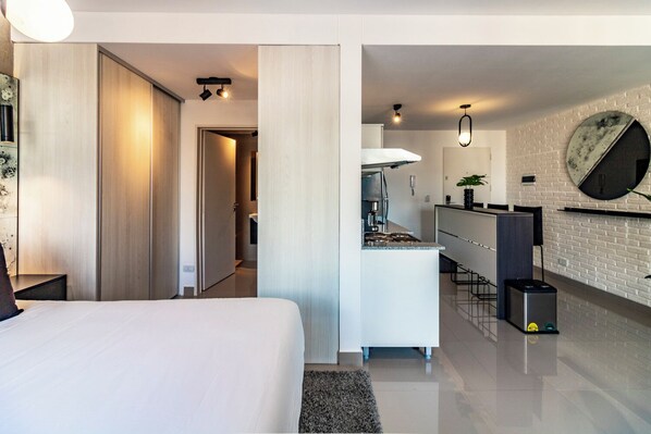 A modern hotel room with a large bed, a kitchenette, and a dining area.