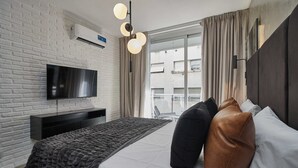 A modern hotel room with a white brick wall, a flat-screen TV, a bed with a grey and white duvet, and a large window with curtains.