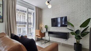 A modern living room with a leather sofa, a desk with a chair, a flat-screen TV mounted on a white brick wall, and a large window with curtains.