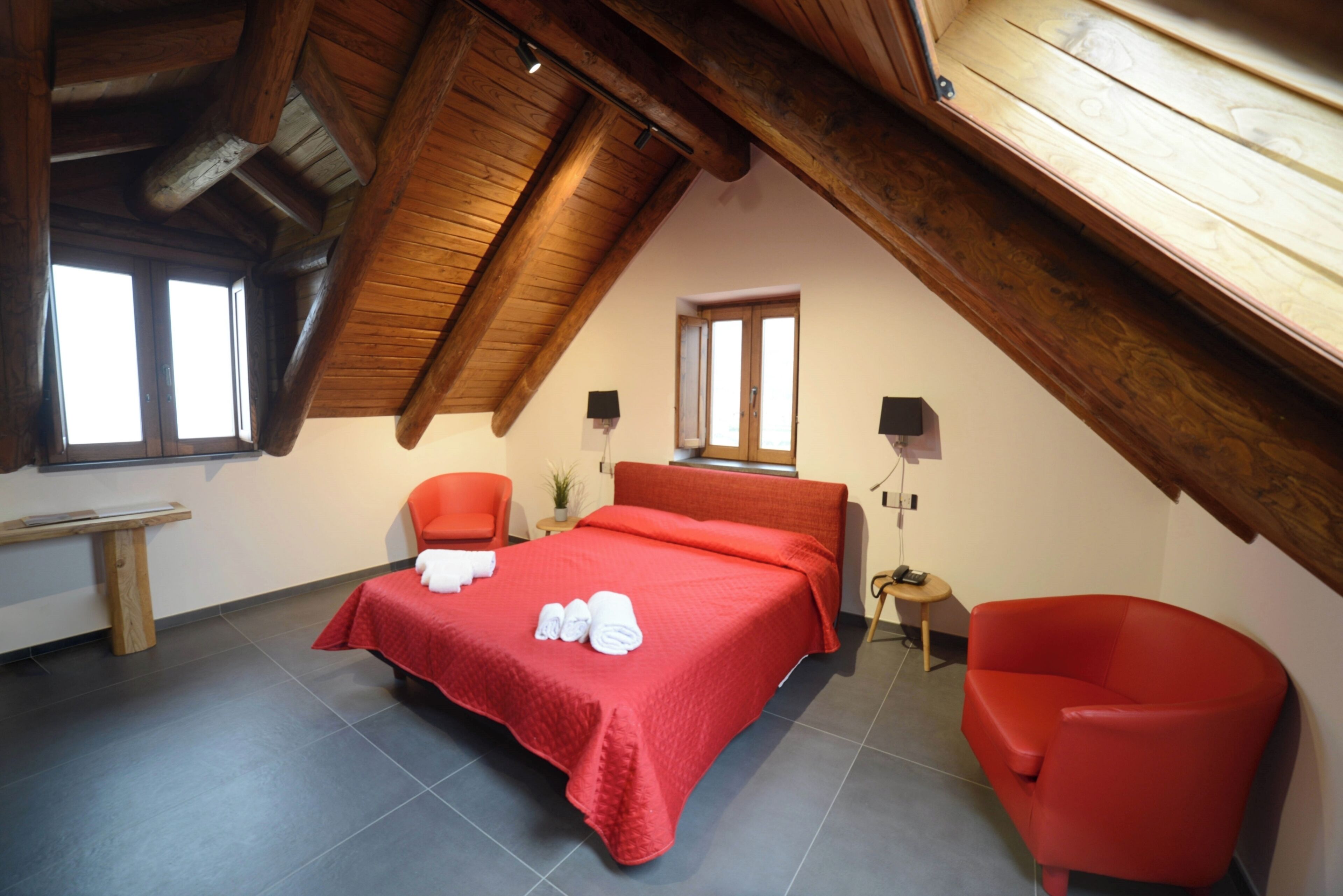 A bedroom with a sloped ceiling, a bed with a red bedspread, two red chairs, and a small table with a lamp.