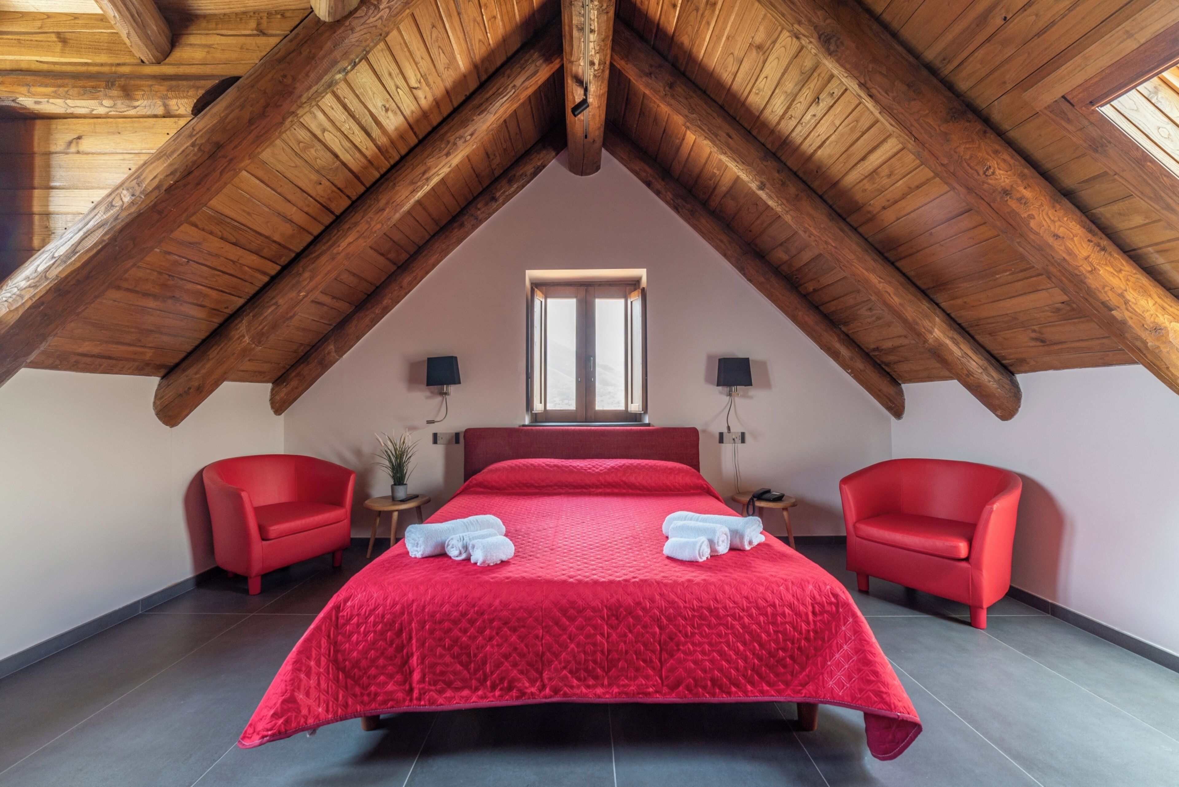 A bedroom with a sloped wooden ceiling, a large bed with red bedding, two red chairs, and a window with natural light.
