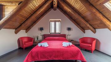 Un dormitorio con techo de madera inclinado, una cama grande con ropa de cama roja, dos sillas rojas y una ventana con luz natural.