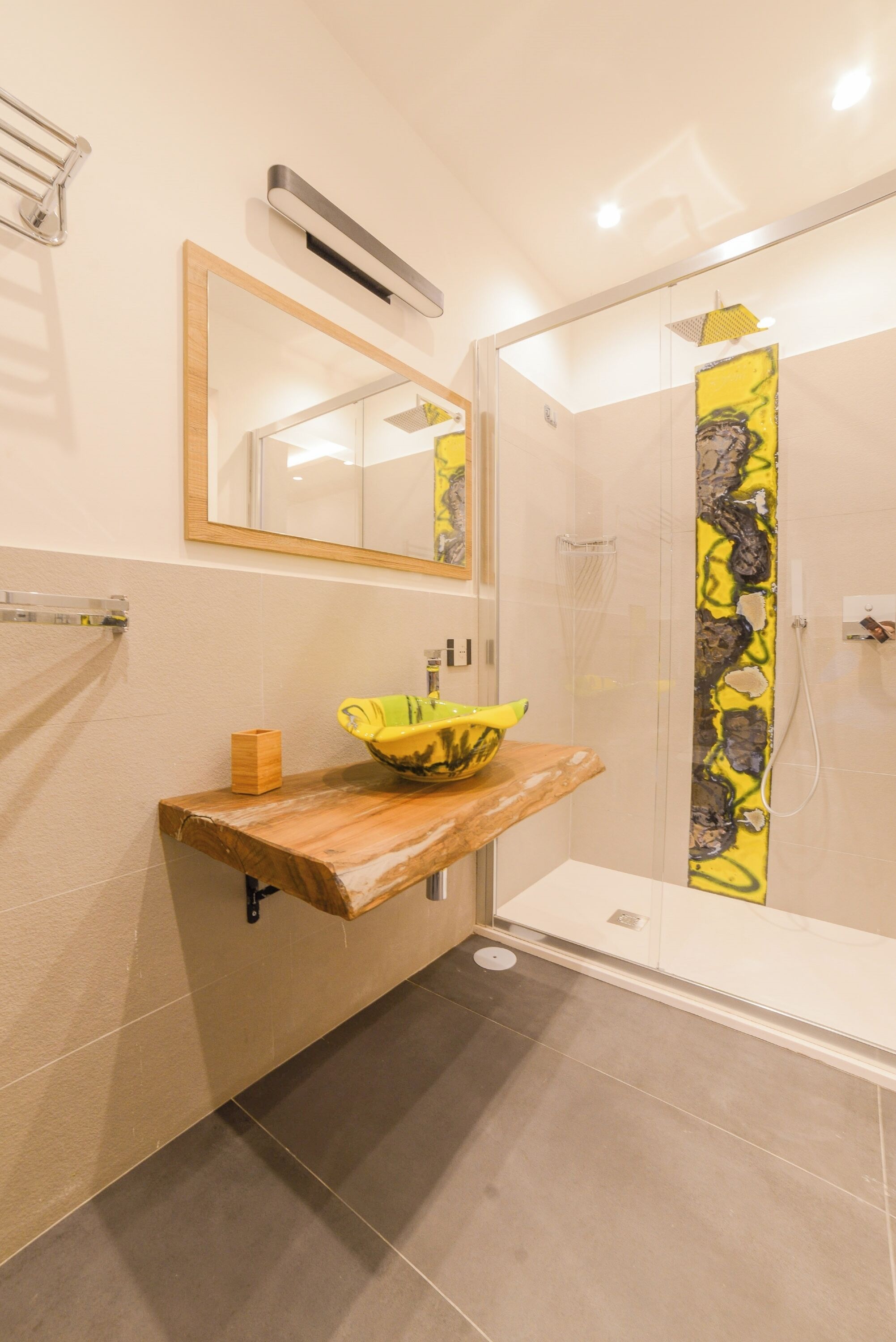 A modern bathroom with a wooden vanity, a yellow sink, a glass shower enclosure, and a mirror above the sink.