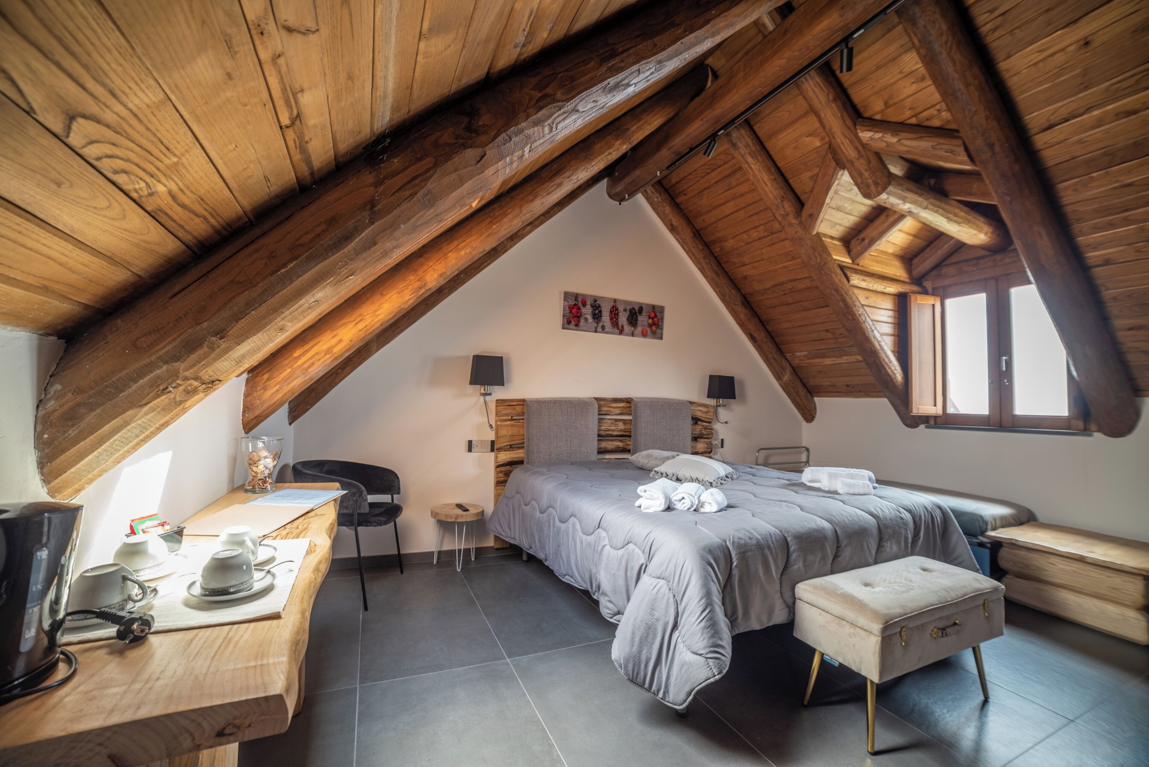 Ein gemütliches Schlafzimmer im Dachgeschoss mit schrägen Wänden, einem Bett mit grauer Bettdecke, einem Holztisch mit Wasserkocher und einem kleinen Hocker.