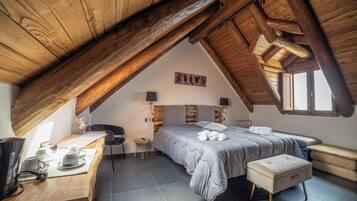 A cozy attic bedroom with a sloped ceiling, a bed with a grey duvet, a wooden desk with a kettle, and a small stool.