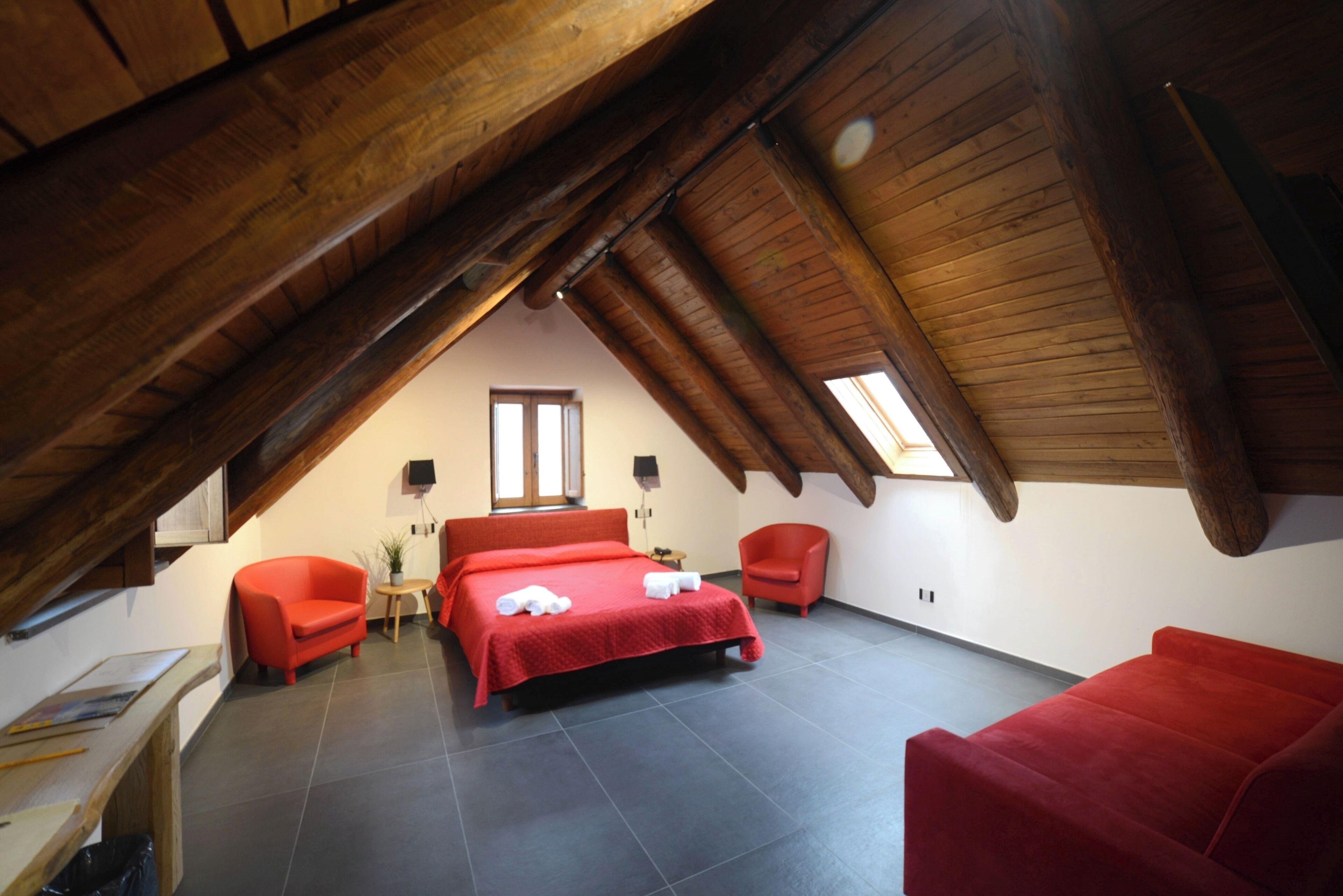 A bedroom with a sloped ceiling, a bed with red bedding, two red chairs, a small table, and a window.