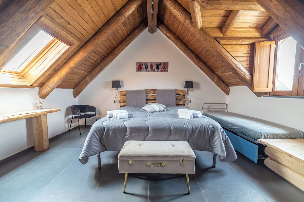 Un dormitorio con techo estilo loft, una cama con edredón gris, un banco de madera, un escritorio y una silla.