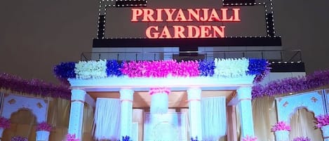 La entrada de Priyanjali Garden, con un letrero iluminado grande y arreglos florales decorativos.
